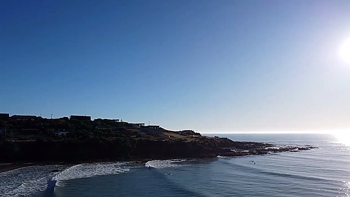 Baches line the headland as a couple of surfers pick off peaks in the glare, North Otago at its quiet best, Oamaru surf spot,
