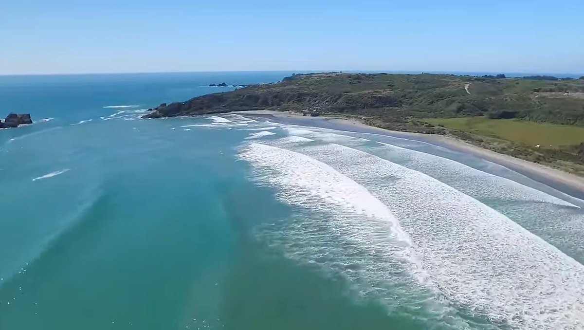 Long lines of whitewater peel down the beach, the bay framed by green farmland and rocky points, Tauranga Bay surf spot, West