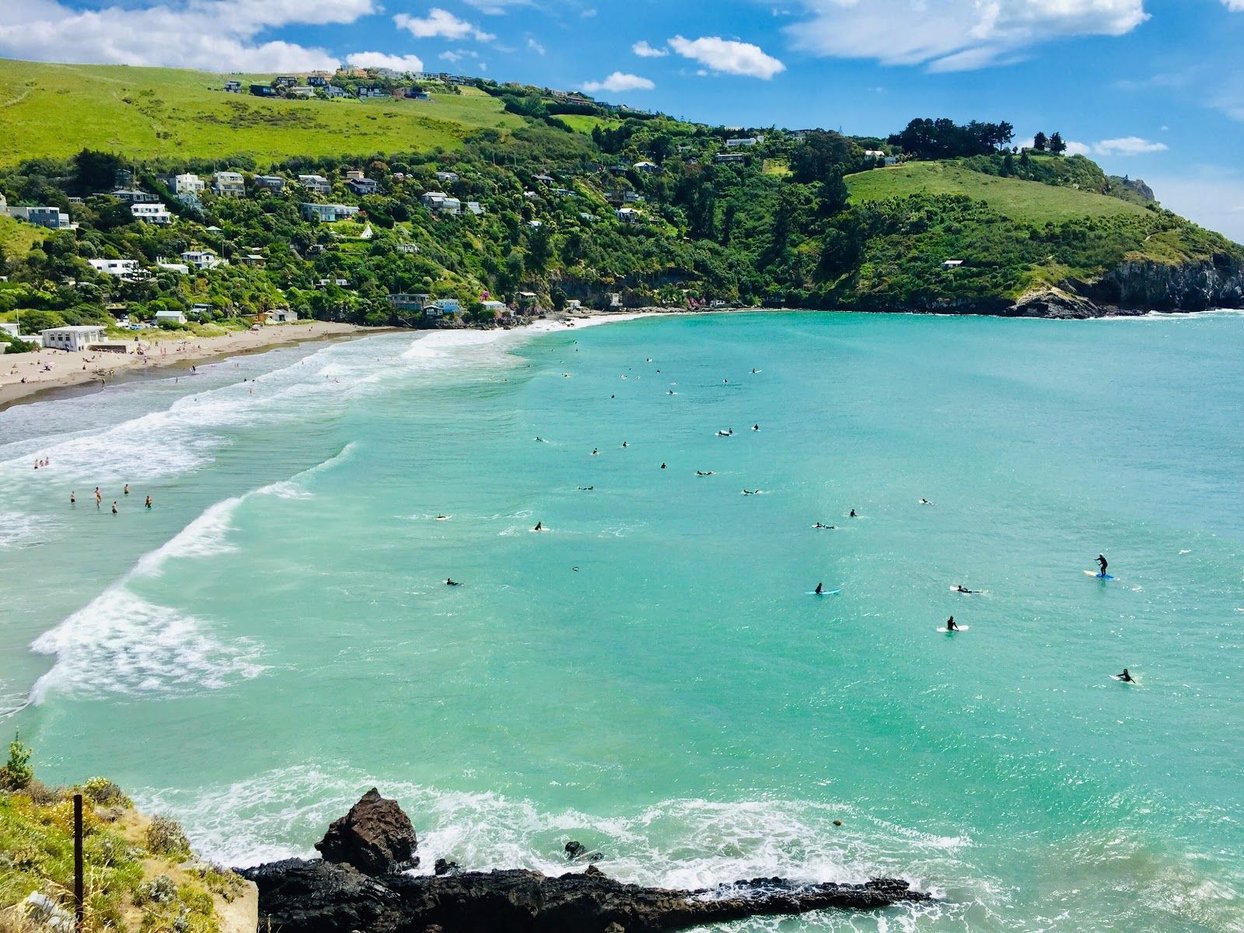 A summer crowd spread across the turquoise bay, baches climbing the green hills above the best wave over the hill from Sumner